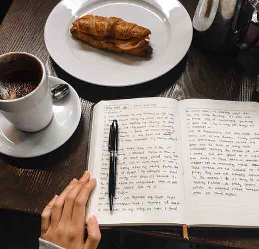 یک میز قهوه‌ای رنگ، که رویش یک بشقاب شکلات داغ و یک بشقاب ساندویچ و یک دفتر ژورنال و خودنویس قرار دارد.